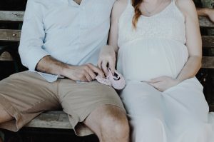 Torso of a pregnant couple, children's shoes on their laps, sitting on a bench.