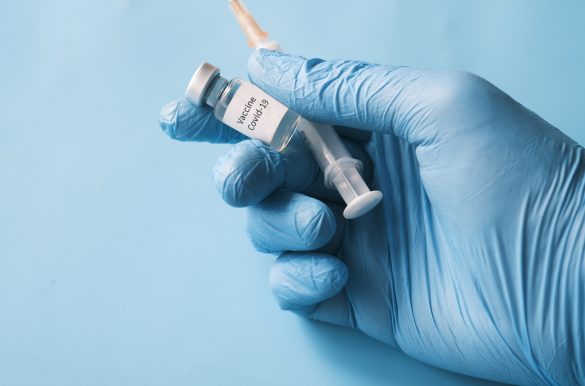 Hand im Latex Handschuh mit COVID-19 Vakzin und Spritze. Beitragsbild zur Charité Kampagne #Impfenschützt, zur 4. Coronawelle.